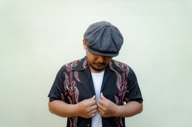 Sakallı ve bıyıklı maço adam bej arka planda çok yalnız görünüyor. Eski moda bir Güneydoğu Asyalı yetişkin adamın yarı vücut portresi desenli gömlek, beyaz atlet bluz ve gazeteci çocuk şapkası.