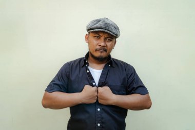 Bıyıklı, sakallı, vahşi maço adamı yakından takip edin. Bej arka planda ciddi ve vahşi bir şekilde izole edilmiş görünüyor. Siyah gömlekli ve şapkalı Güneydoğu Asyalı bir yetişkinin yarı vücut portresi.