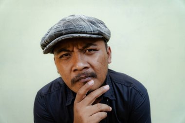 Bıyıklı, sakallı, vahşi maço adamı yakından takip edin. Bej arka planda ciddi ve vahşi bir şekilde izole edilmiş görünüyor. Siyah gömlekli ve şapkalı Güneydoğu Asyalı bir yetişkinin yarı vücut portresi.