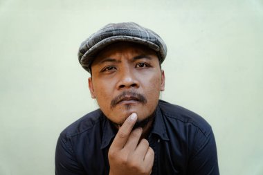 Bıyıklı, sakallı, vahşi maço adamı yakından takip edin. Bej arka planda ciddi ve vahşi bir şekilde izole edilmiş görünüyor. Siyah gömlekli ve şapkalı Güneydoğu Asyalı bir yetişkinin yarı vücut portresi.