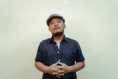 Bıyıklı, sakallı, vahşi maço adamı yakından takip edin. Bej arka planda ciddi ve vahşi bir şekilde izole edilmiş görünüyor. Siyah gömlekli ve şapkalı Güneydoğu Asyalı bir yetişkinin yarı vücut portresi.