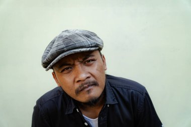 Bıyıklı, sakallı, vahşi maço adamı yakından takip edin. Bej arka planda ciddi ve vahşi bir şekilde izole edilmiş görünüyor. Siyah gömlekli ve şapkalı Güneydoğu Asyalı bir yetişkinin yarı vücut portresi.