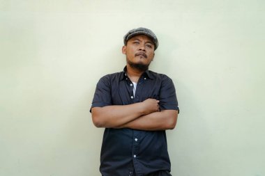 Bıyıklı, sakallı, vahşi maço adamı yakından takip edin. Bej arka planda ciddi ve vahşi bir şekilde izole edilmiş görünüyor. Siyah gömlekli ve şapkalı Güneydoğu Asyalı bir yetişkinin yarı vücut portresi.