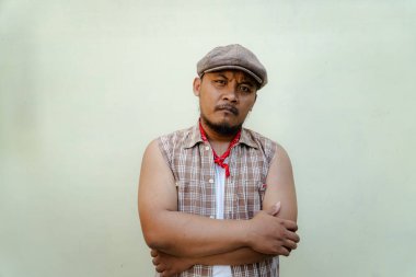 Southeast Asian macho man with beard and mustache looks serious and fierce isolated on beige background. Half body portrait of adult man in sleeveless shirt, bandana and newsboy hat