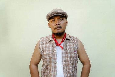 Southeast Asian macho man with beard and mustache looks serious and fierce isolated on beige background. Half body portrait of adult man in sleeveless shirt, bandana and newsboy hat