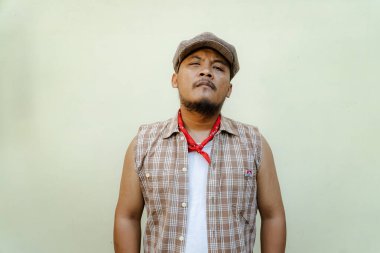 Southeast Asian macho man with beard and mustache looks serious and fierce isolated on beige background. Half body portrait of adult man in sleeveless shirt, bandana and newsboy hat