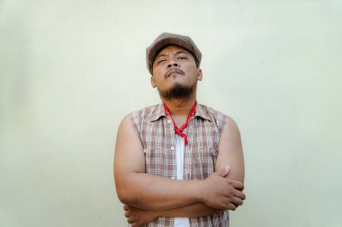 Southeast Asian macho man with beard and mustache looks serious and fierce isolated on beige background. Half body portrait of adult man in sleeveless shirt, bandana and newsboy hat