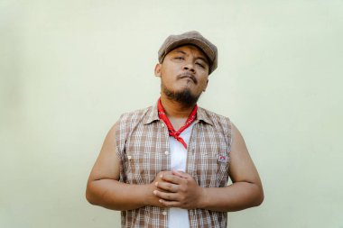 Southeast Asian macho man with beard and mustache looks serious and fierce isolated on beige background. Half body portrait of adult man in sleeveless shirt, bandana and newsboy hat