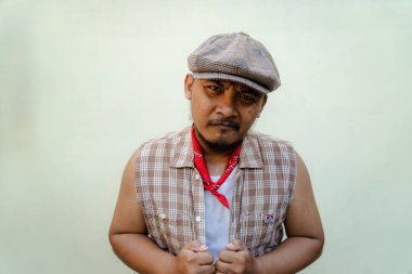 Southeast Asian macho man with beard and mustache looks serious and fierce isolated on beige background. Half body portrait of adult man in sleeveless shirt, bandana and newsboy hat