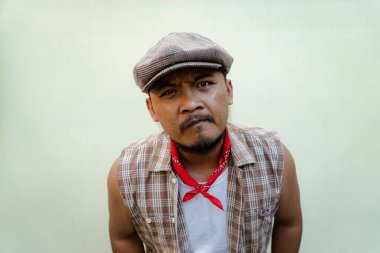 Southeast Asian macho man with beard and mustache looks serious and fierce isolated on beige background. Half body portrait of adult man in sleeveless shirt, bandana and newsboy hat