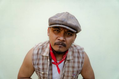 Southeast Asian macho man with beard and mustache looks serious and fierce isolated on beige background. Half body portrait of adult man in sleeveless shirt, bandana and newsboy hat