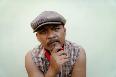 Southeast Asian macho man with beard and mustache looks serious and fierce isolated on beige background. Half body portrait of adult man in sleeveless shirt, bandana and newsboy hat