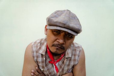 Southeast Asian macho man with beard and mustache looks serious and fierce isolated on beige background. Half body portrait of adult man in sleeveless shirt, bandana and newsboy hat