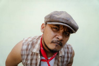 Southeast Asian macho man with beard and mustache looks serious and fierce isolated on beige background. Half body portrait of adult man in sleeveless shirt, bandana and newsboy hat