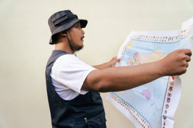 Asian adult man wearing travel outfit is looking at world map on beige background. Half body portrait of adult Southeast Asian man posing wearing vest and boonie hat reading a map clipart