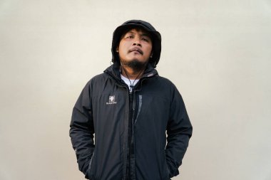 Sakallı ve bıyıklı maço bir yetişkin adam ciddi poz veriyor ve krem rengi bir arka plana hafifçe gülümsüyor. Yetişkin bir Güneydoğu Asyalı 'nın yarı vücut portresi şapka, tişört ve ceket giyiyor.