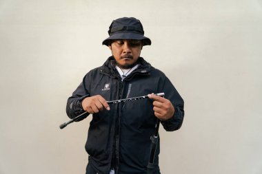 Portrait of Asian man wearing hiking outfit. Half body portrait of adult Asian man with beard and mustache posing climber wearing jacket, boonie hat and trekking poles isolated on beige background.