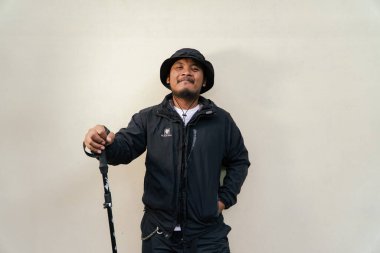 Portrait of Asian man wearing hiking outfit. Half body portrait of adult Asian man with beard and mustache posing climber wearing jacket, boonie hat and trekking poles isolated on beige background. clipart