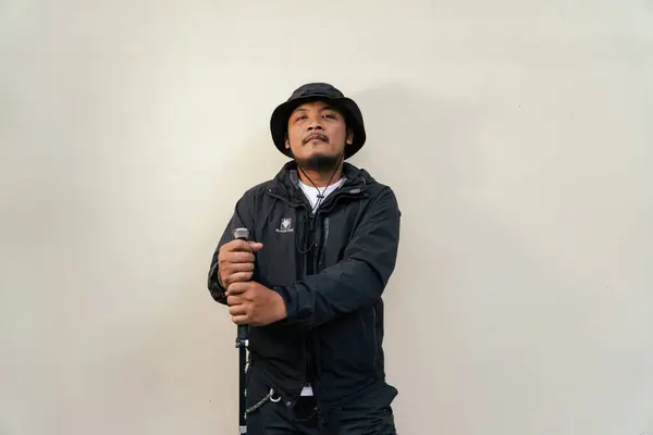Portrait of Asian man wearing hiking outfit. Half body portrait of adult Asian man with beard and mustache posing climber wearing jacket, boonie hat and trekking poles isolated on beige background.