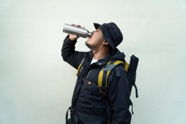 Ceketli, şapkalı, sırt çantalı ve bej arka planda izole edilmiş içki şişesi giyen bir adam. Şişeden su içen yetişkin bir Asyalının yarı vücut portresi.