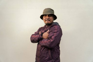 Half body portrait of mature Asian man with beard and mustache posing in ready style before traveling. Mature man wearing adventure outfit with jacket and bucket hat isolated on beige background.