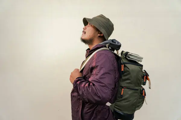 Half body portrait of adult Asian man with beard and mustache posing and wearing hiking outfit with jacket, bucket hat, carrier bag and trekking poles isolated on beige background.