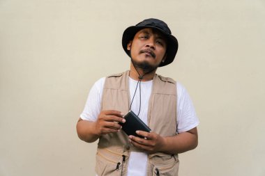 Mature asian man wearing travel outfit with vest, bucket hat and hip flask bottle isolated on beige background. Half body portrait of an adult Asian man posing drinking from a hip flask bottle clipart