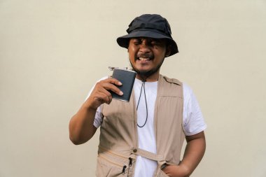 Mature asian man wearing travel outfit with vest, bucket hat and hip flask bottle isolated on beige background. Half body portrait of an adult Asian man posing drinking from a hip flask bottle clipart