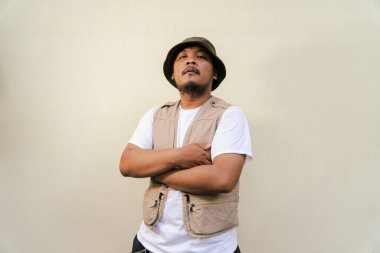Macho mature man with beard and mustache wearing safari clothes isolated on beige background. Half body portrait of an adult Southeast Asian man striking various poses wearing a vest and bucket hat clipart
