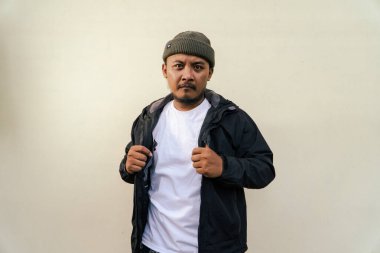 A macho mature man with a beard and mustache poses seriously and fiercely against a beige background. Half body portrait of adult Southeast Asian man posing wearing beanie cap, t-shirt and jacket