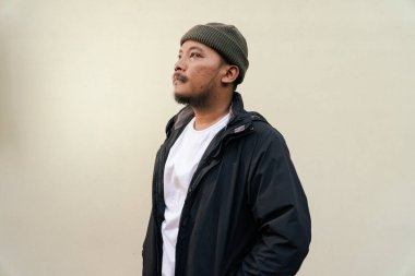 A macho mature man with a beard and mustache poses seriously and fiercely against a beige background. Half body portrait of adult Southeast Asian man posing wearing beanie cap, t-shirt and jacket clipart