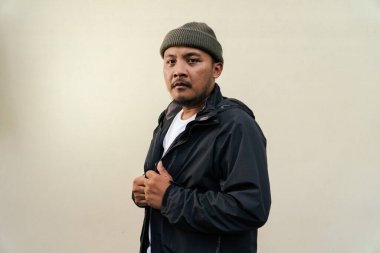 A macho mature man with a beard and mustache poses seriously and fiercely against a beige background. Half body portrait of adult Southeast Asian man posing wearing beanie cap, t-shirt and jacket
