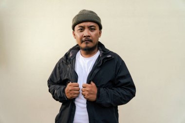 A macho mature man with a beard and mustache poses seriously and fiercely against a beige background. Half body portrait of adult Southeast Asian man posing wearing beanie cap, t-shirt and jacket