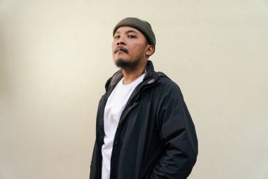A macho mature man with a beard and mustache poses seriously and fiercely against a beige background. Half body portrait of adult Southeast Asian man posing wearing beanie cap, t-shirt and jacket