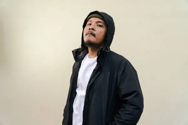 A macho mature man with a beard and mustache poses seriously and fiercely against a beige background. Half body portrait of adult Southeast Asian man posing wearing beanie cap, t-shirt and jacket