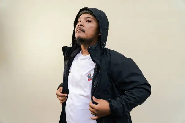 A macho mature man with a beard and mustache poses seriously and fiercely against a beige background. Half body portrait of adult Southeast Asian man posing wearing beanie cap, t-shirt and jacket