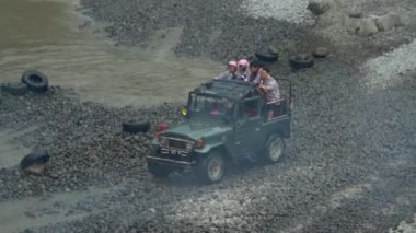 Bir off-road turist cipinin 4K görüntüsü yüksek hızda nehri geçerken su tüm yönlere sıçrayıp yolcuları Merapi Dağı eteklerindeki lav turunda ıslatırken görülüyor.