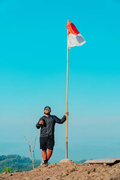 stock image A man at the top of the ancient volcano Nglanggeran which is dominated by rocks and has the red and white Indonesian flag. Photo of adventurer reaching rocky mountain peak with flag waving