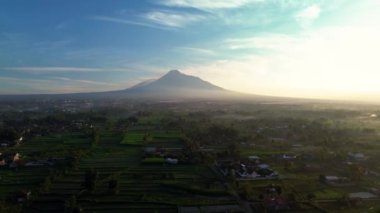 Endonezya, Yogyakarta 'daki Merapi Dağı' nın 4k hava görüntüsü. Güneşin doğuşundan kısa bir süre sonra sabah manzarası... pirinç tarlaları ve yerleşim alanları ön planda.