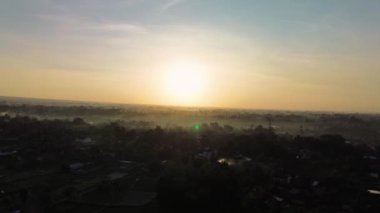 Etrafı ağaçlarla ve sislerle dolu bir köyde baş döndürücü bir altın güneş doğuşunun 4K hava görüntüsü. Güneşin hala güzel ve gür olan kırsal bir bölgede doğduğu an.