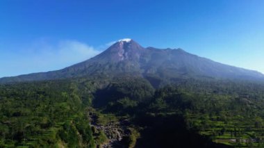 Merapi Dağı 'nın panoramik manzaralı, güzel bir yaz sabahı manzaralı hava aracı uçuş videosu. Önünde yeşil bir orman olan etkin bir volkanın 4k manzarası.