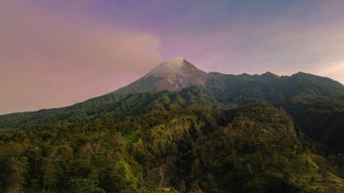 Güneş doğarken Merapi Dağı 'nın manzarası çok güzeldir. Önünde mor bir gökyüzü, ormanı ve vadisi olan aktif bir volkanın detaylı manzarası.
