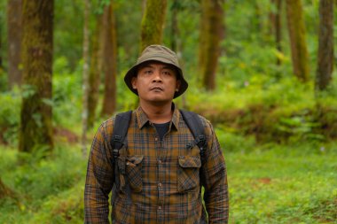 Yoğun ve yeşil bir çam ormanında bir maceracının yarı vücut görüntüsü. Flanel gömlekli, kova şapkalı ve sırt çantalı Asyalı bir erkek gezgin yoğun çam ağaçlarının arkasında poz veriyor..