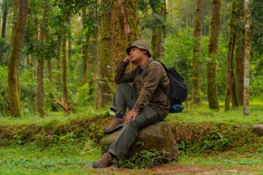 Yoğun bir çam ormanında büyük bir kayanın üzerinde oturan bir maceraperest. Flanel gömlekli ve şapkalı Asyalı bir adam oturmuş güzel doğanın huzurunun tadını çıkarırken dinleniyor..