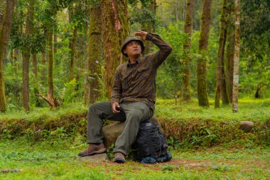 Yoğun bir çam ormanında büyük bir kayanın üzerinde oturan bir maceraperest. Flanel gömlekli ve şapkalı Asyalı bir adam oturmuş güzel doğanın huzurunun tadını çıkarırken dinleniyor..