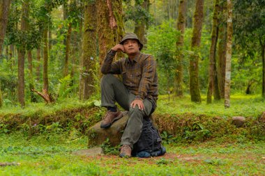 Yoğun bir çam ormanında büyük bir kayanın üzerinde oturan bir maceraperest. Flanel gömlekli ve şapkalı Asyalı bir adam oturmuş güzel doğanın huzurunun tadını çıkarırken dinleniyor..