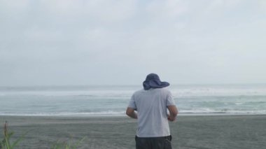 Rüzgarlı bir plajın kenarında kumsalda yürüyen yalnız bir maceracının görüntüsü. Bir erkek gezgin kumsalda tek başına yürüyor.