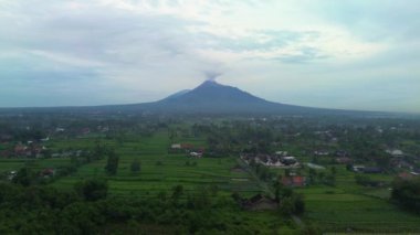 Güneş uzaktan yükselirken Merapi Dağı 'nın panoramik güzelliğinin 4K hava görüntüsü. Sabahları dağın havadan görünüşü kırsal ve pirinç tarlalarının panoramik görüntüsüyle.