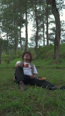 Çam ormanlarının yemyeşil bölgesinde kızartılmış tempeh atıştırması yiyen bir adamın 4K görüntüsü. Asyalı maceracının çimenlerde oturduğu ve sakin bir atmosferde yemek yerken büyük bir kayaya yaslandığı yakın çekim görüntüsü.