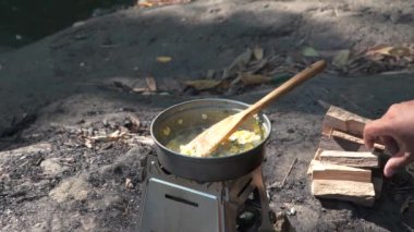 Bushcraft konsepti basit bir odun sobası kullanarak nehir kenarında sandviç ve sıcak çay hazırlıyor. Erkek bir maceracı, yemyeşil ağaçlarla çevrili temiz bir nehrin kenarında kum cadıları ve kaynar su pişiriyor.